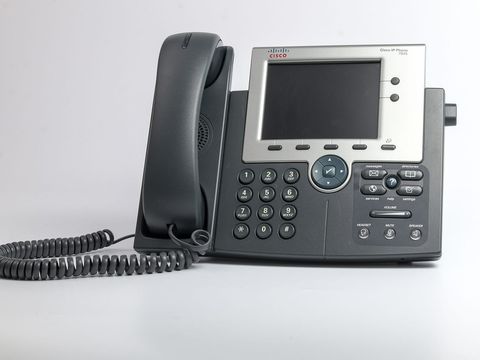 Graues IP-Tischtelefon mit Farbdisplay und Spiralkabel vor hellem Hintergrund