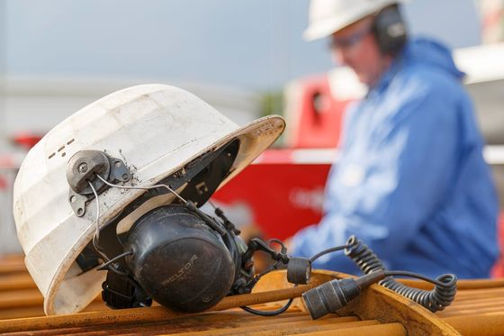 Weißer Schutzhelm mit Gehörschutz-Headset auf Stahlrohren, im Hintergrund ein Arbeiter