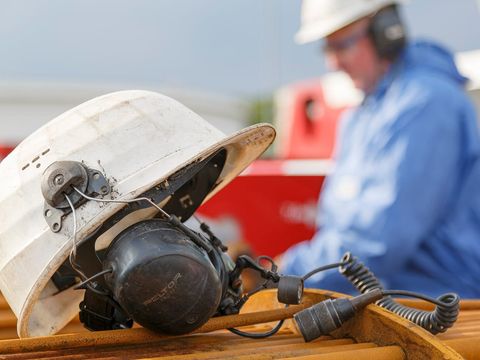 Weißer Schutzhelm mit Gehörschutz-Headset auf Stahlrohren, im Hintergrund ein Arbeiter