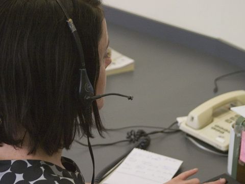 Frau mit Headset am Schreibtisch, daneben Tischtelefon und handschriftliche Notizen, Blick von hinten