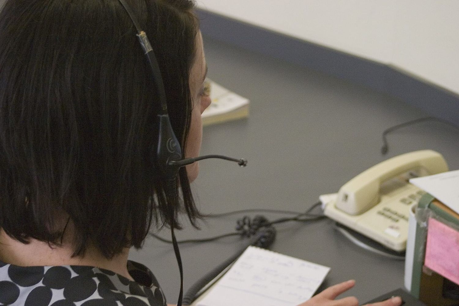 Frau mit Headset am Schreibtisch, daneben Tischtelefon und handschriftliche Notizen, Blick von hinten